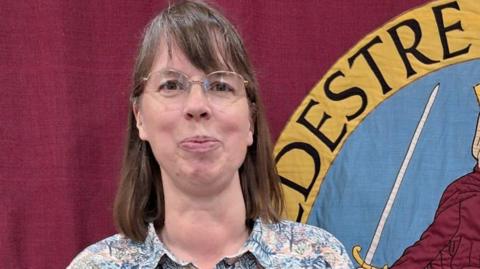 A woman with shoulder length brown hair and wire-framed glasses stands in front of a large town flag in maroon with part of a yellow, black and blue crest visible.