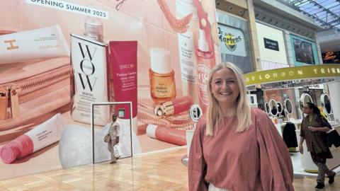 A blonde woman wearing a pink shirt smiling inside of a shopping centre 