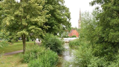 A river running through a park underneath a small white bridge. Surrounded by trees and mowed grass, with a church spire and houses visible in the background