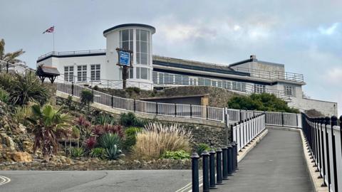 An exterior view of the art deco Winter Gardens building. It is a white building constructed of various shapes, with dirt on some walls. It sits on the cliff edge with a long path leading up to it.