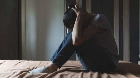 A woman with dark hair, a grey T-shirt and jeans sits on a bed with her head in her hands