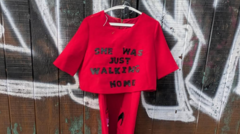 A small red top embroidered with black letters reading "she was just walking home" hanging outside as part of the Raising the Red project. 