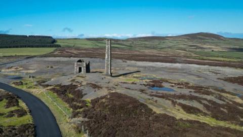 Drone-shot image of Cross Vein mine. The original chimney and out-house can be seen surrounded by wild headland and gravel. A road passes through the field.