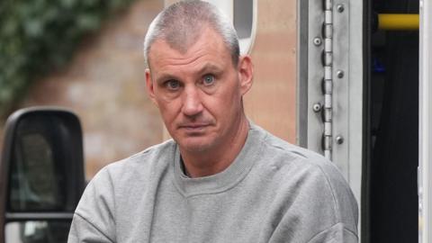 A man with short grey hair wearing a grey tracksuit top. He is being taken out of a prison van and staring at the camera.