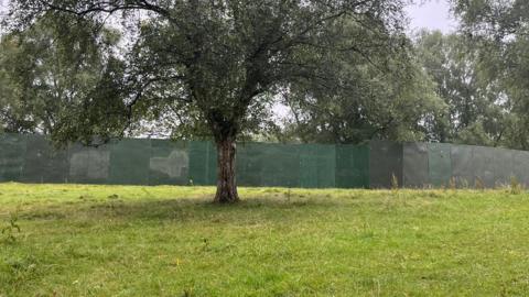 A large green fence erected across a green field with tree seen in the foreground. 