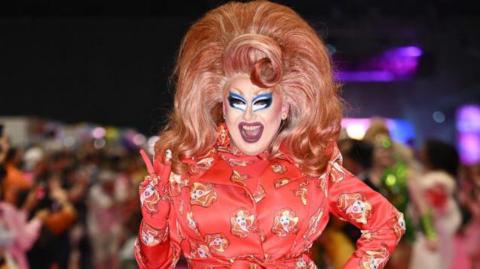 Drag artist Porkchop with a red outfit on and wearing a large red wig with blonde streaks.