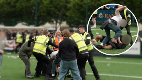 Marshalls wearing hi-viz vests attempt to separate brawling fans on a football pitch
