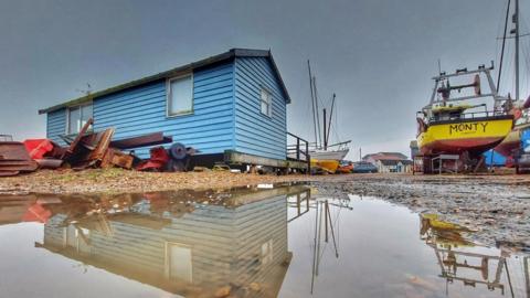 A blue boat house and a yellow boat are in front of a huge puddle with grey sky behind