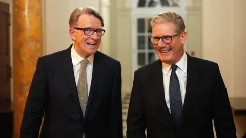Mandelson and Starmer - both wearing suits - walk down a hallway together, smiling.