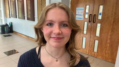 Kaitlyn. She has long loght brown hair, wearing a navy v-neck jumper and necklace.