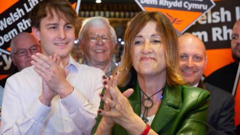 Jane Dodds at the launch event surrounded by Lib Dem campaigners. Her hands are together and outstretched in front of her. She is wearing and wearing a shiny green jacket. There are orange Liberal Democrat banner behind her.
