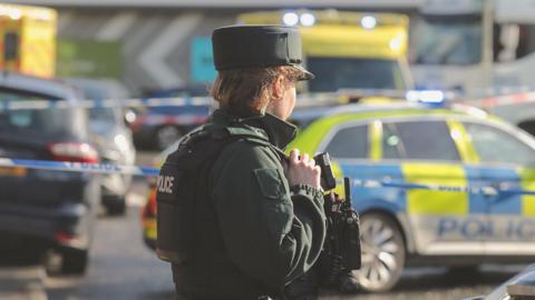 A female police officer stands with her back to the camera. In the distance is a police car, which is parked in front of some police tape.