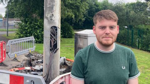 A man in a green shirt stands in front of a metal lamp post. There is a hole cut out of the post with thick wiring protruding from the hole, it is scorched and badly damaged. 