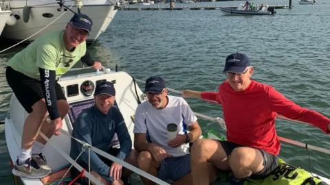 A photo of four men sat on a small sailboat. Two are sat on the floor, one on a bag which is on the floor, and one is holding onto the edges of the boat and standing balanced on one side. The boat is docked at the time, with other boats of different sized on the background. It is a sunny day, and all four men are wearing caps, and smiling towards the camera.