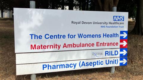 A hospital sign reads Royal Devon University Healthcare and directs people to The Centre for Womens Health, Maternity Ambulance Entrance as well as the  RILD (Research Innovation Learning & Development) building and Pharmacy Aseptic Unit.