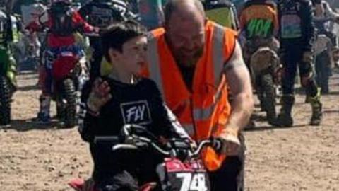 A young boy in a black long-sleeved shirt sits on a motocross bike on a muddy track. A man, with a bald head and ginger beard and wearing a sleeveless high-vis jacket crouches next to him.