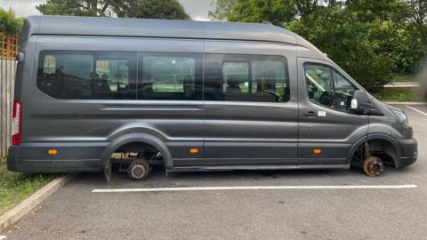 A dark grey Ford Transit van resting on the ground of a car park with its axles exposed having had its tyres stolen.