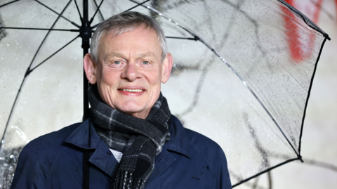 Martin Clunes smiles for the cameras as he arrives for the UK Premiere of 'Wuthering Heights' at Odeon Luxe Leicester Square in London. He has short grey hair, is holding a large clear umbrella and is wearing a navy blue coat and black tartan scarf.