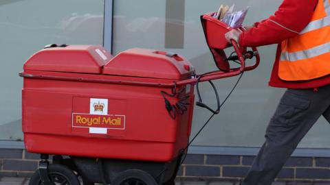 A picture of a worker for Royal Mail wearing a red top and orange jacket. He is pushing a red Royal Mail trolley. There are letters on top of it.