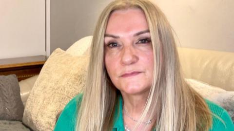 A woman with blonde hair, wearing a green top, sat on a sofa and looking into the camera