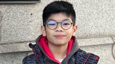 A young boy looking directly in the camera. He has glasses and dark hair.