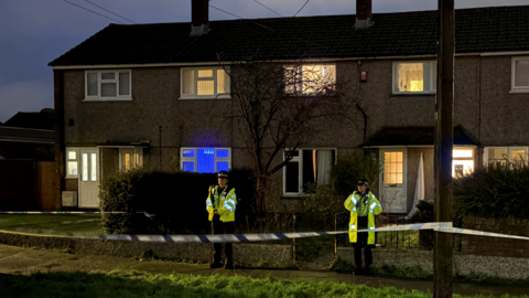 Two police officers in high-visibility clothing stand behind a cordon at night. There are three terrace houses behind them. Most of the windows are lit up by bright lights but some have curtains and blinds drawn.