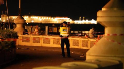 A policeman stands next to a cordoned off area on Brighton seafront with his back to the camera. It is night-time and the area is bathed in an orange glow from streetlights. Bright lights from seaside attractions can be seen in front of the police officer. A man is sitting on a wall with his back to the officer and a man and woman walk past, looking at the officer as they do so.