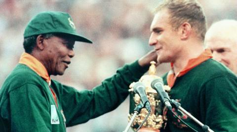 Nelson Mandela, wearing a green cap and predominantly green Springboks jersey, goes to tap Francois Pienaar on the left shoulder as the South Africa captain holds the Rugby World Cup trophy. Two microphones on stands are seen in the foreground of the picture