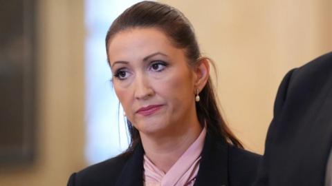 Emma Little-Pengelly tilts her head to the right. She has long dark hair tied back. She looks stern. She is wearing a pink neck scarf and black jacket.