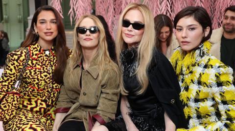 Dua Lipa, Vanessa Paradis, Nicole Kidman and Gracie Abrams sit together on a pink seat at a fashion show, with pink weeping willows behind them - Paradis and Kidman wear dark glasses and hold hands, smiling, while Abrams wears a bright yellow tweed jacket, and Lipa also wears yellow, patterned with black and red