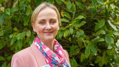 Rachel Street stands in front of a hedge. She is smiling and wearing a pink blazer and a multicoloured scarf. She has short blonde hair.