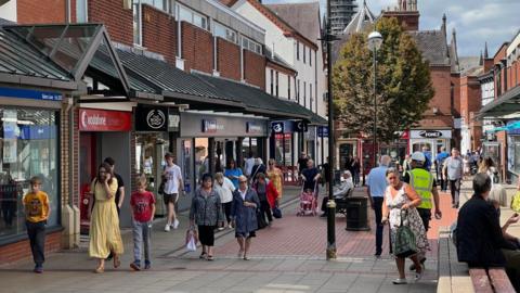 A city centre high street filled with people walking along it. There is a row of shops on the left and lamp-posts and a tree in the middle of the street.