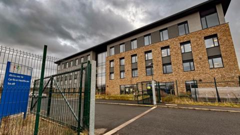 Side view of a large four storey building - a health centre - with rows of windows and a glassed-in area in the centre. There is wire fencing around it, with a large open gate for the access road. A sign to the left reads: Welcome to Carlisle Healthcare South