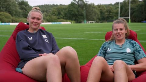 Two women's rugby players sit on bean bags on the pitch and smile at the camera
