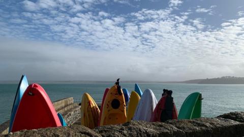 Multi-coloured surf boards on end by a wall with the blue sea and blue sky behind