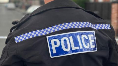 A stock image of a police officer pictured from behind. In the head-and-shoulders image he is wearing a black uniform jacket with the police logo on the back and a uniform hat. He is standing on a residential street where a police tape is cordoning off an area. The background is blurred.
