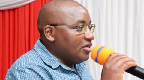 George Koimburi, in a checked light blue and white shirt, holding a microphone 