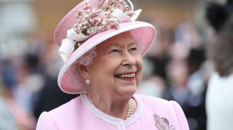 Queen Elizabeth smiling and wearing a pink outfit, pink hat and pearl necklace