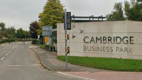 The entrance to the Cambridge Business Park. On the right is a stone wall with Cambridge Business Park written on it in silver letters. There is a traffic light in front of it. The road stretches down and has small trees. 