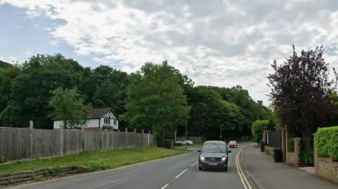 A leafy road with cars driving on it and a white house in the background.