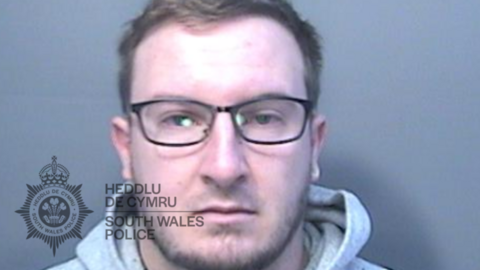 A mugshot of a man with short fair hair and dark glasses. he is wearing a grey hoodie and looking straight at the camera with a neutral expression.