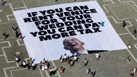 A large banner against Amazon founder Jeff Bezos lies on the ground, placed by Greenpeace Italy activists and UK activist group Everyone Hates Elon, on the ground of St Mark's Square, on 23 June, 2025. It is white with black lettering in capitals and has a picture of Bezos throwing his head back laughing
