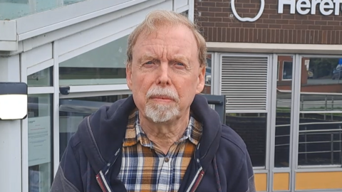 Councillor Louis Stark, a man with light brow hair who is wearing a yellow checked shirt with a navy blue jacket. He is standing outside a council building with glass entrance and railings.