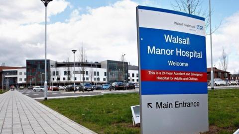 Walsall Manor Hospital in the distance - a white and grey building with a car park in front of it. There is a hospital sign in the foreground pointing people to the main entrance.