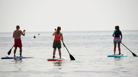 Two women and one man are standing up on paddleboards with oars in their hands. The man is wearing only shorts, the woman in the middle is wearing a vest top and shorts and the woman to the right is wearing a rash vest and shorts.