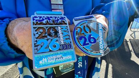 Two medals are held up by a man wearing a blue coat. One medal is sqaure and reads "Coventry Half Marathon" while the second is round and reads "Coventry 10k". 