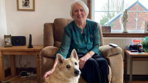 Di Stirling has a grey/platinum-blonde short bob hairstyle and is wearing a dark green top. She is sitting in an armchair in her living room with her dog Sirius sitting at her feet looking towards the camera. Di is smiling at the camera