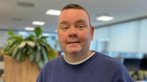 A man in a blue jumper stands in an office building.