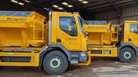 Three yellow gritter lorry. They have the word 'Cormac' on the side on door and large gritters on the back.