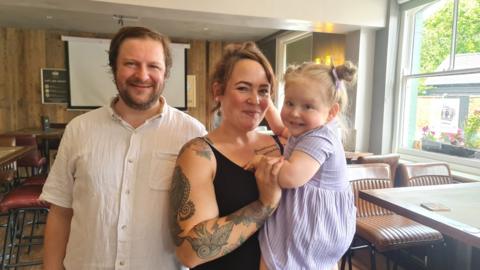 A man in a white shirt next to a woman with a black dress holding a child in a purple dress